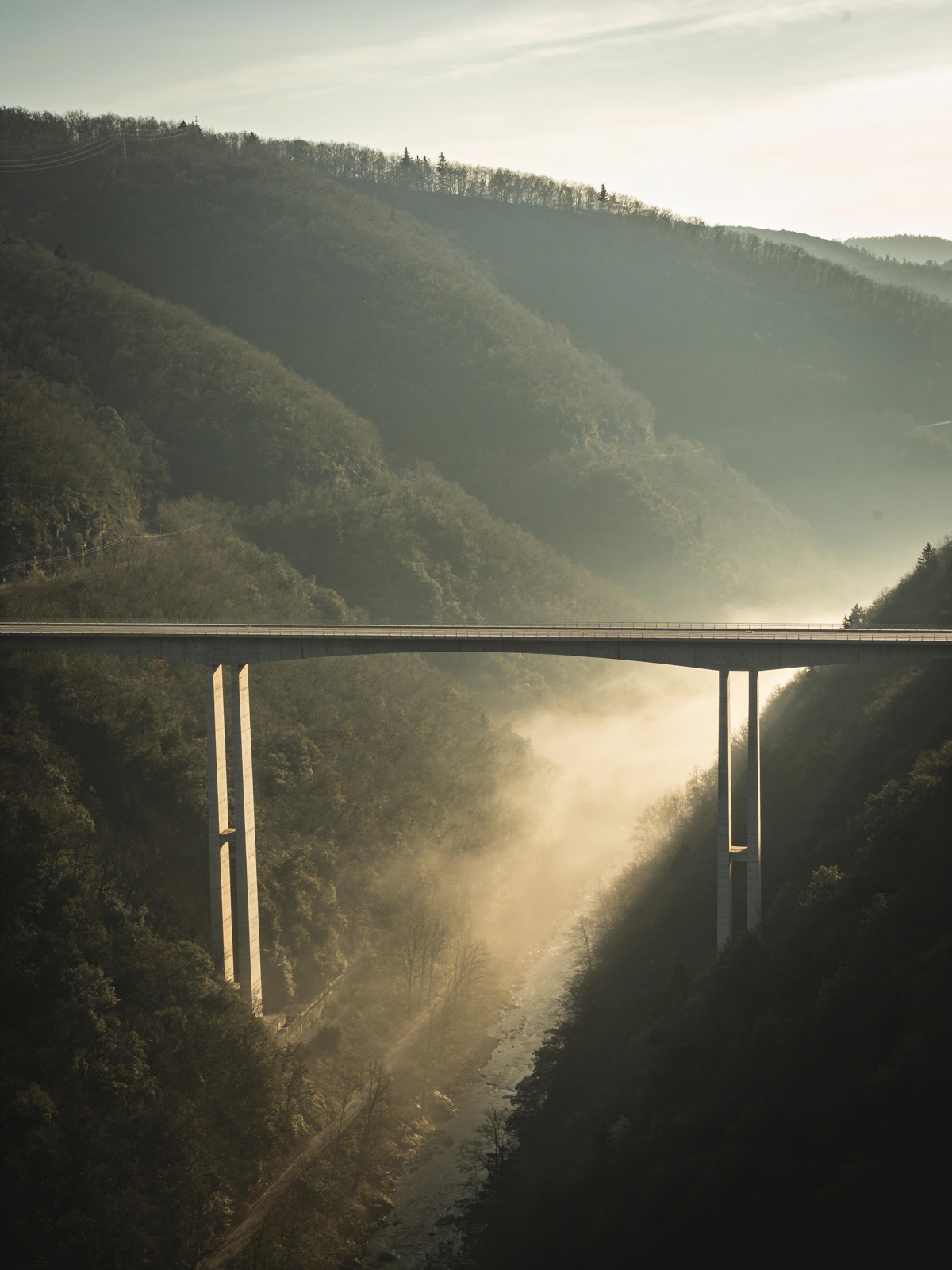 A long concrete bridge spanning a misty river valley, both of its ends anchored on opposite slopes.