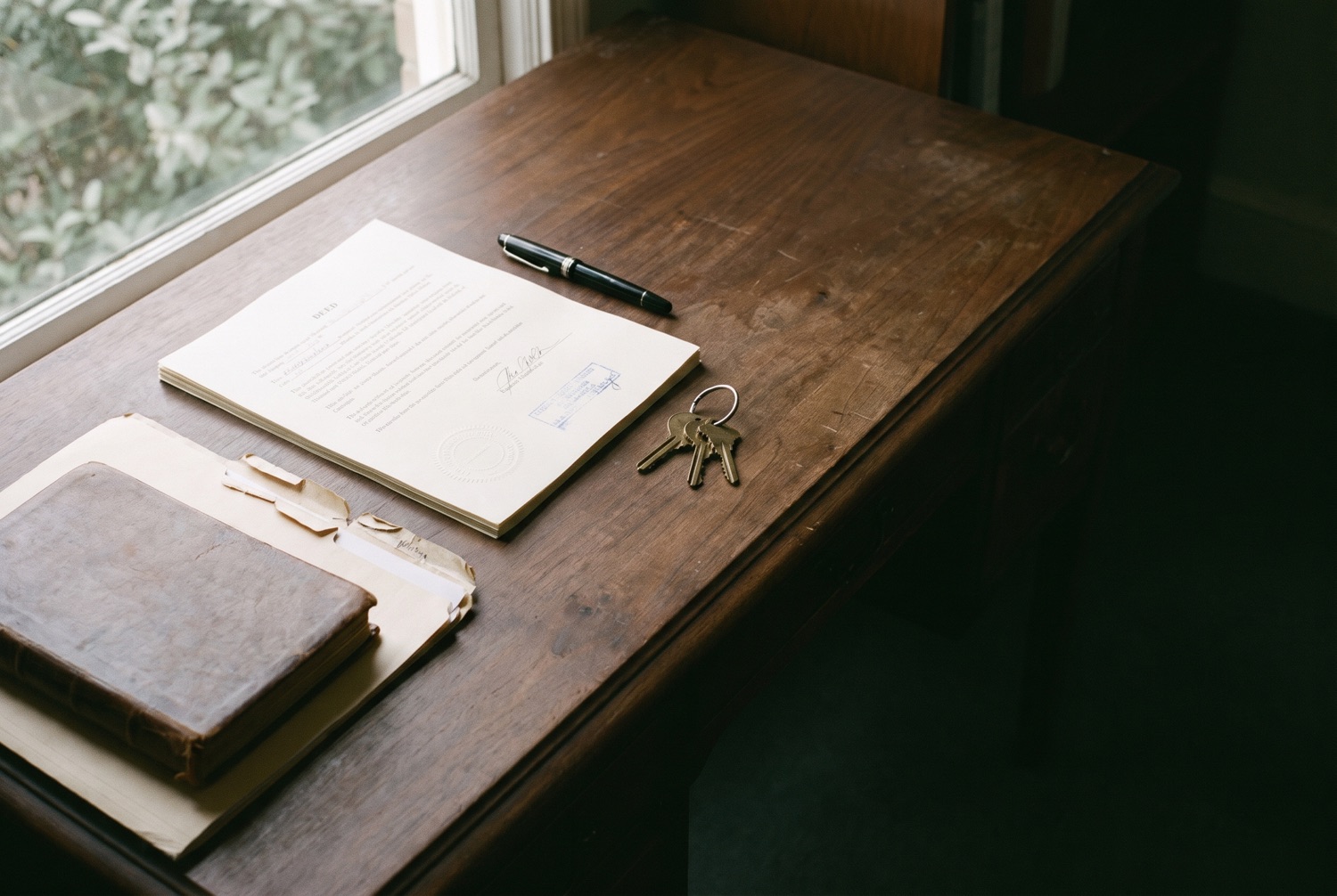 A notary's desk after a completed closing: a signed deed with an embossed seal, a capped fountain pen, house keys and a leather-bound protocol book.