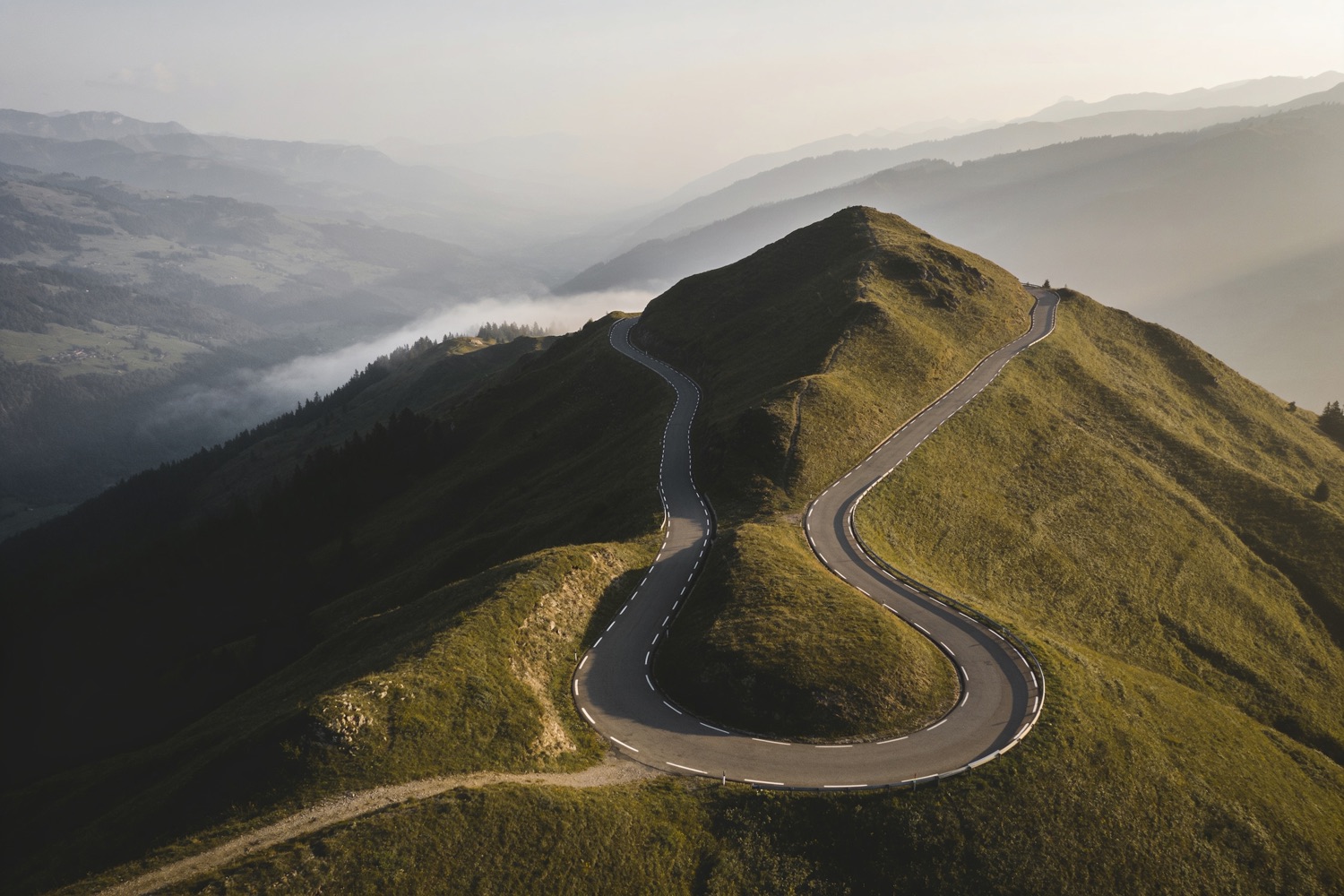 A single road winding over a mountain pass at first light, seen from above.